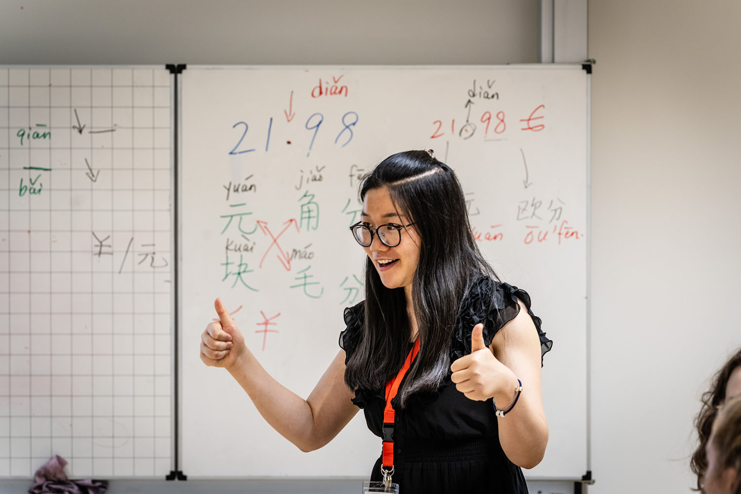Eine junge Frau mit asiatischen Gesichtszügen steht vor einer mit chinesischen Schriftzeichen beschriebenen Tafel und hebt lächelnd beide Daumen. 