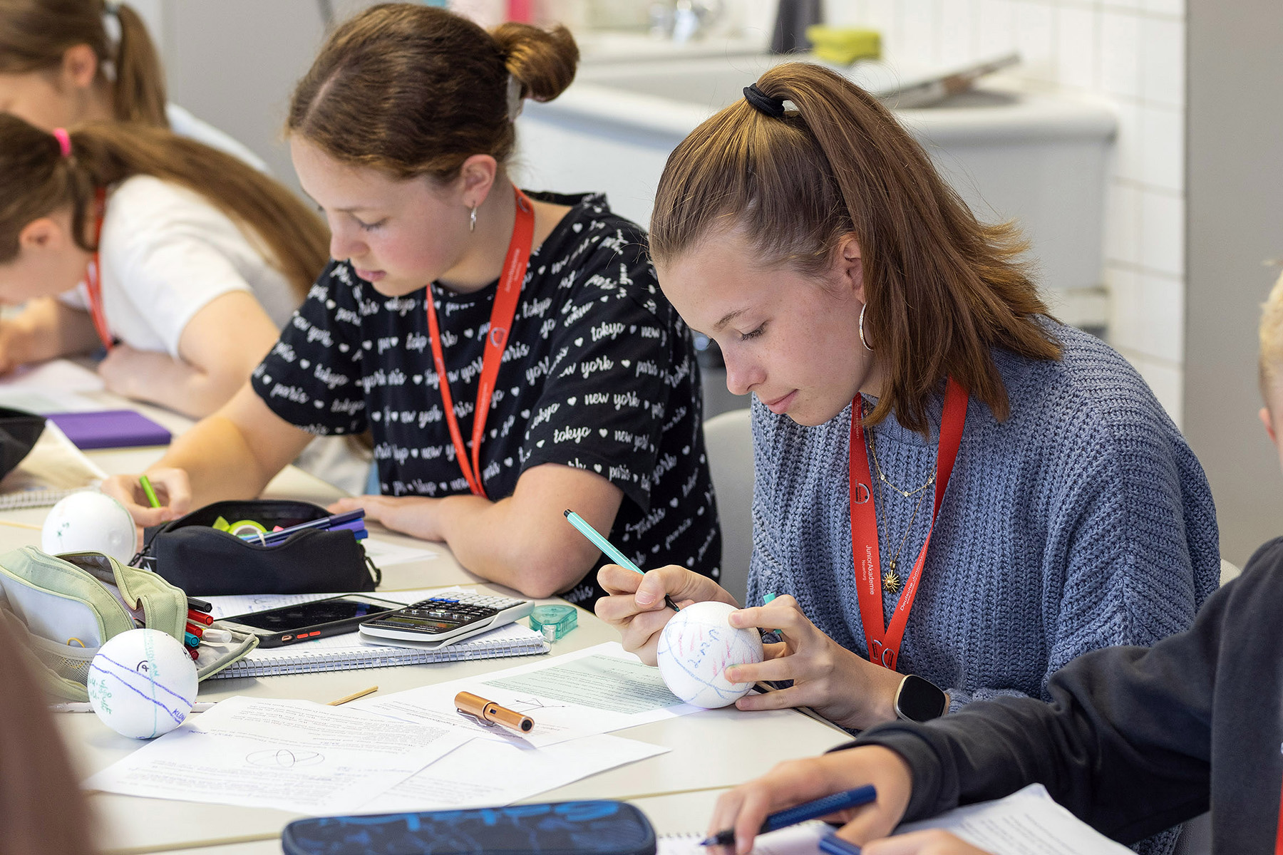 Zwei junge Menschen arbeiten konzentriert an kleinen Kunststoffkugeln, die sie mit Formeln beschriften. Auf dem Tisch vor ihnen liegt viel Arbeitsmaterial.