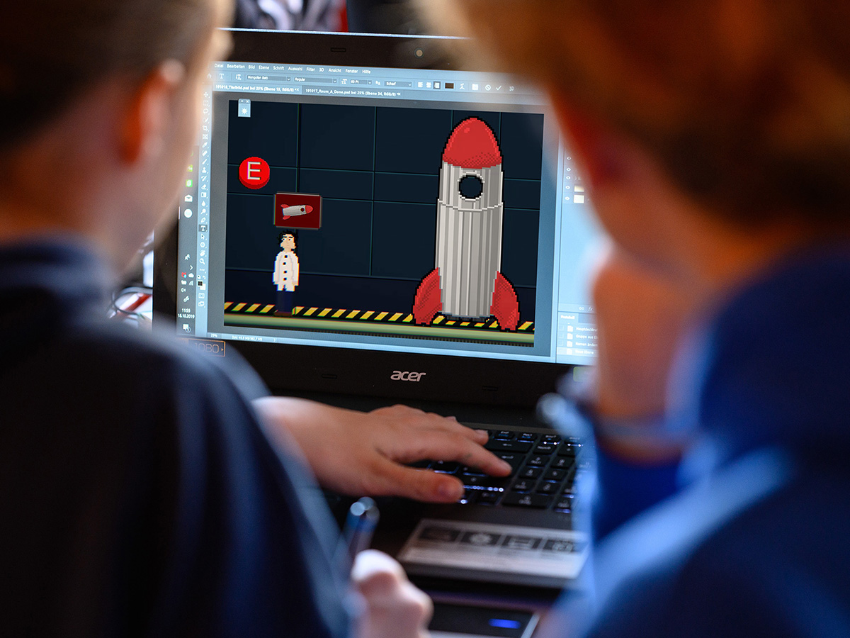 Von hinten: Zwei Jugendliche arbeiten konzentriert an einem Laptop, auf dem ein Bild in Pixelgrafik zu sehen ist.
