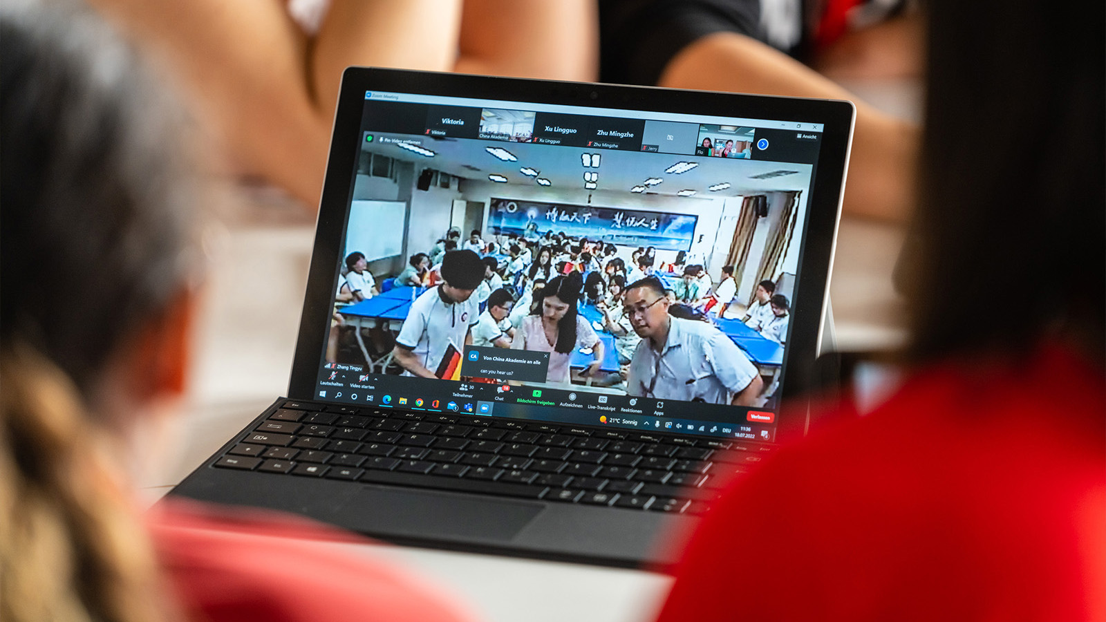 Zwei Jugendliche sitzen vor einem aufgeklappten Laptop, auf dem man eine Gruppe chinesischer Schülerinnen und Schüler erkennt.