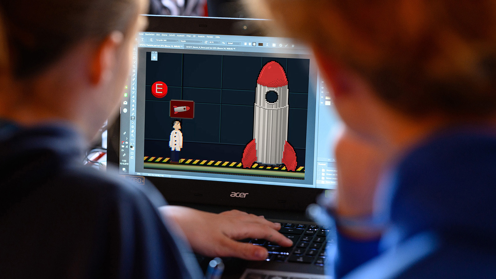 Von hinten: Zwei Jugendliche arbeiten konzentriert an einem Laptop, auf dem ein Bild in Pixelgrafik zu sehen ist.