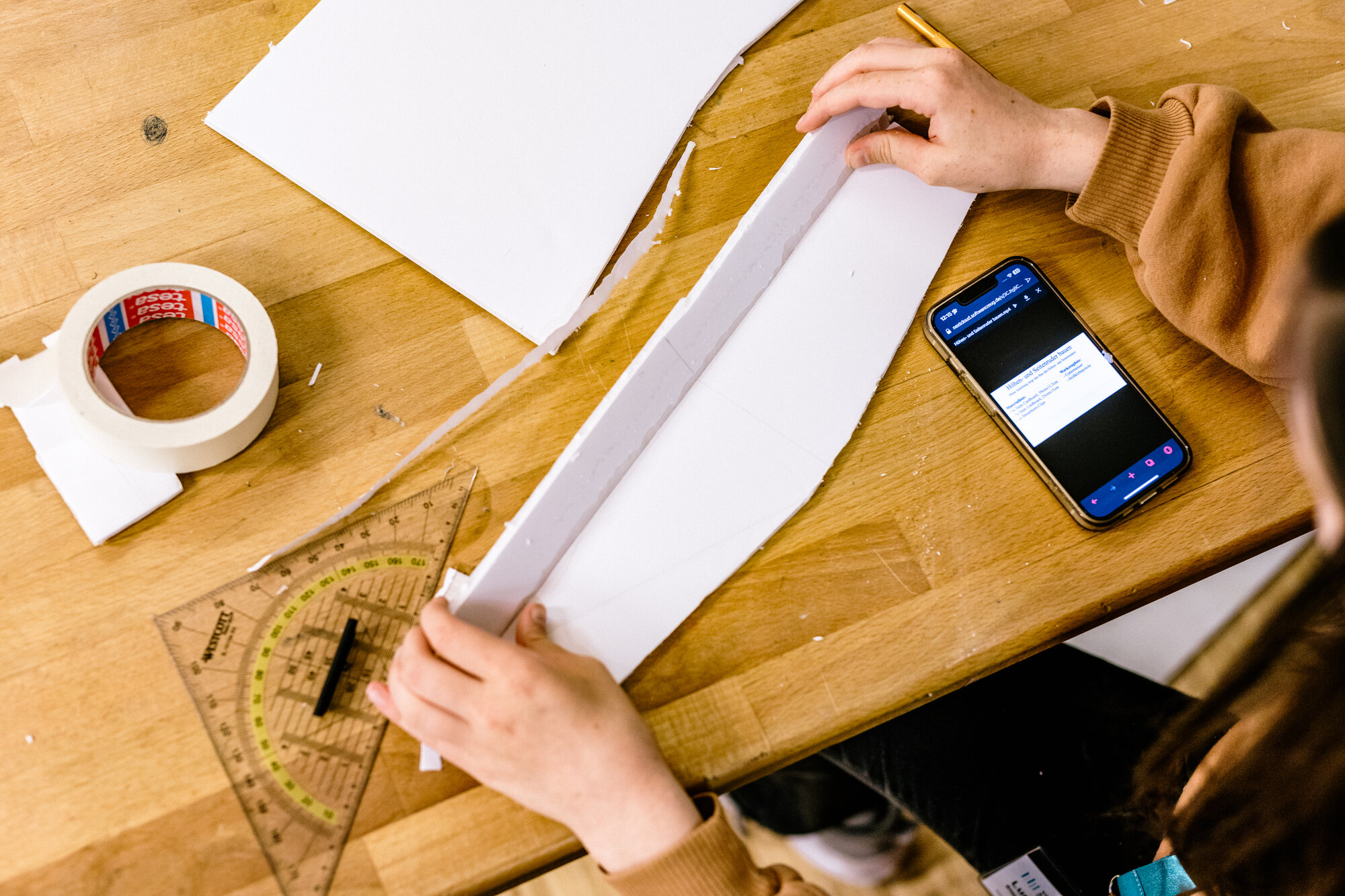 Draufsicht auf zwei Hände, die ein Blatt Papier falten. Daneben ein Handy, ein Geodreieck und eine Rolle mit Tesafilm. 