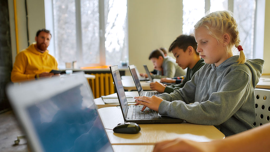 Junge Schülerinnen und Schüler sitzen mit einem Lehrer in einem Klassenraum und arbeiten an Laptops.