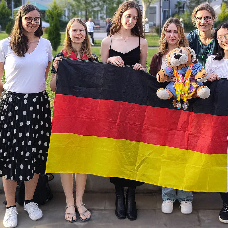 Die vier Teilnehmerinnen an der EGMO 2024 zusammen mit den beiden Delegationsleiterinnen, der deutschen Flaggs und dem Maskottchen "MathemaTigerin", der mehrere Medaillen umgehängt wurden. 