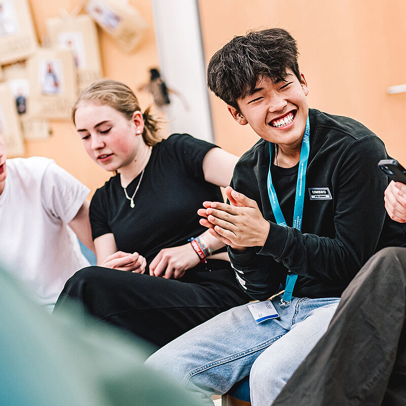 Vier Jugendliche sitzen in einer Stuhlreihe, einer von ihnen lächelt in die Kamera.