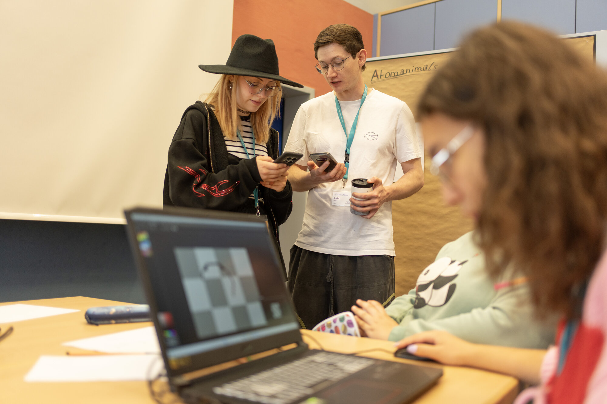 Zwei kreativ gekleidete Jugendliche die in einem Klassenraum stehen und sich mit ihren Smartphone abstimmen. Im Vordergrund arbeitet eine Jugendliche am Laptop an einer Grafik.